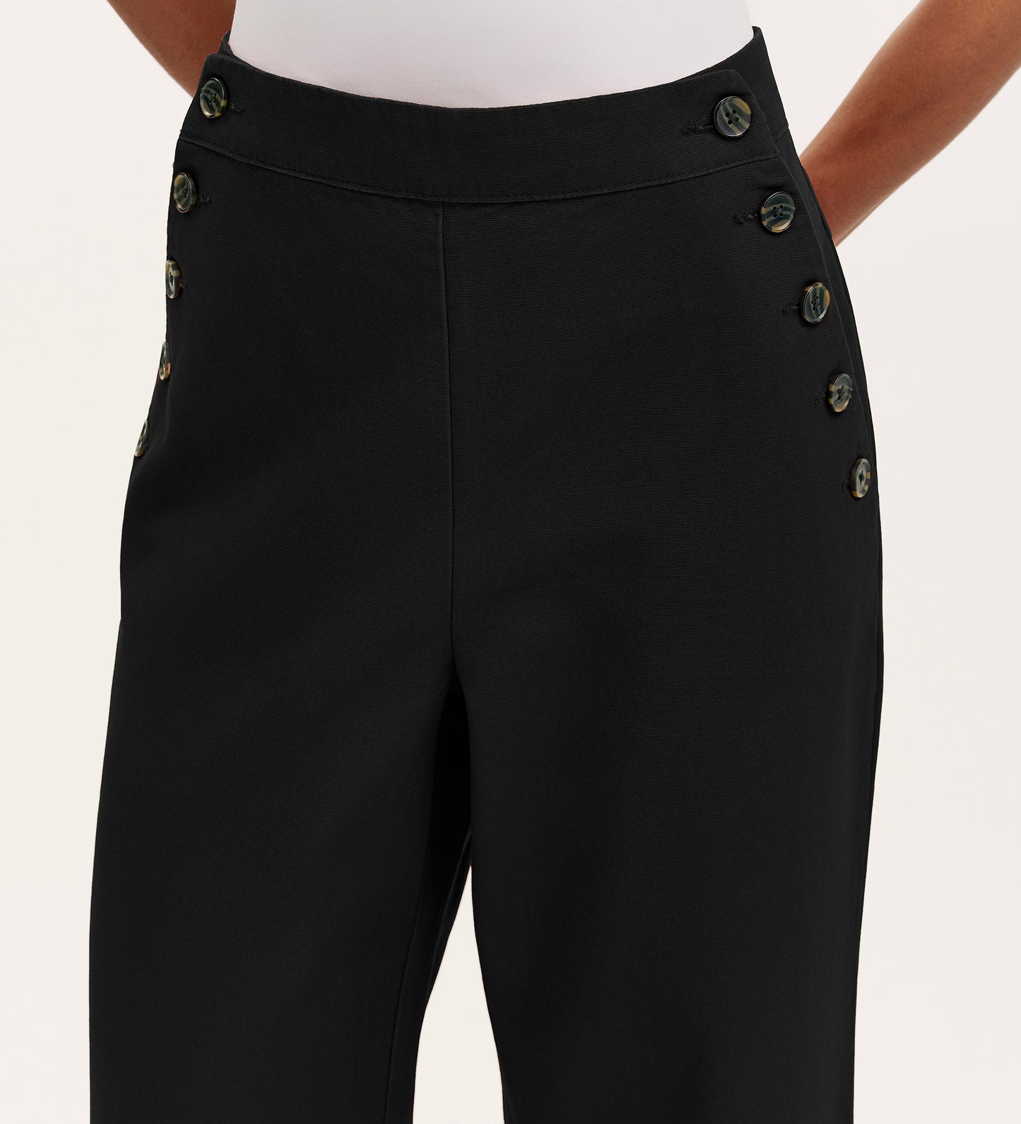 A person wears Nellie Black Canvas Wide Leg Trousers featuring decorative buttons along the waistband, paired with a white top. The image is cropped just below the chest.