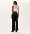 A woman faces away, wearing a white sleeveless top, heeled shoes, and the Nellie Black Canvas Wide Leg Trousers—a versatile wardrobe staple—against a plain, light-colored background.