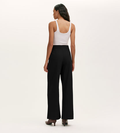 A woman faces away, wearing a white sleeveless top, heeled shoes, and the Nellie Black Canvas Wide Leg Trousers—a versatile wardrobe staple—against a plain, light-colored background.