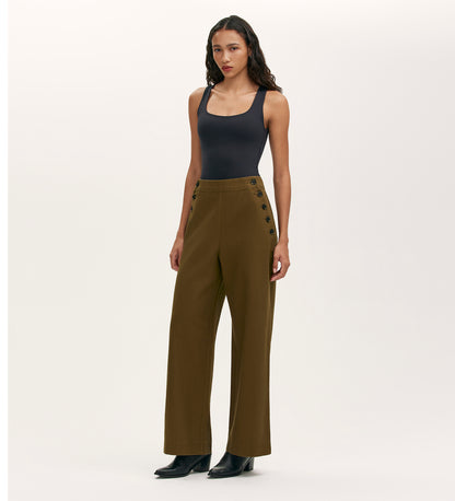 A person stands against a plain white background wearing a fitted black tank top, the Nellie Khaki Canvas Wide Leg Trousers with side buttons—an essential piece—paired with black boots.