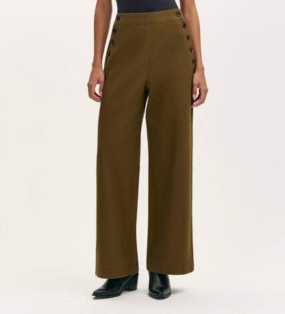 A person wears Nellie Khaki Canvas Wide Leg Trousers with double rows of black buttons, paired with a black top and black heeled ankle boots. The background is plain and light-colored.