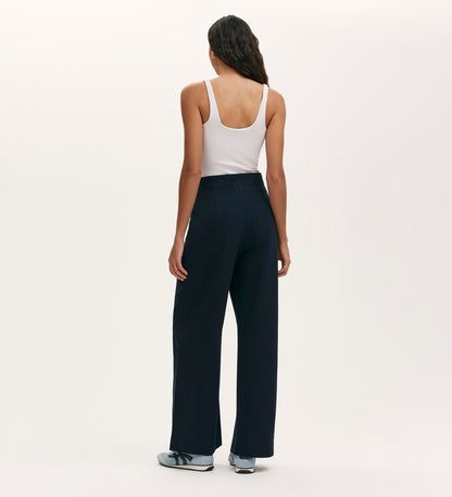 A person with long dark hair, seen from behind against a plain light background, wears a white tank top, light blue sneakers, and the Nellie Navy Canvas Wide Leg Trousers—a true wardrobe staple.