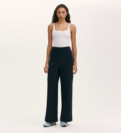 A woman faces forward against a plain light background wearing a white tank top, Nellie Navy Canvas Wide Leg Trousers with side button details, and light blue sneakers.