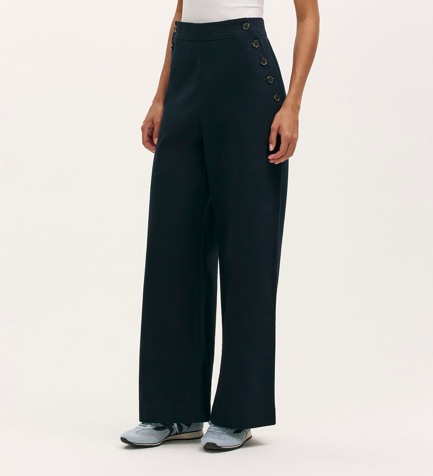 A person in Nellie Navy Canvas Wide Leg Trousers with side button details, a white sleeveless top, and gray sneakers stands against a plain light background—a timeless choice for modern style.
