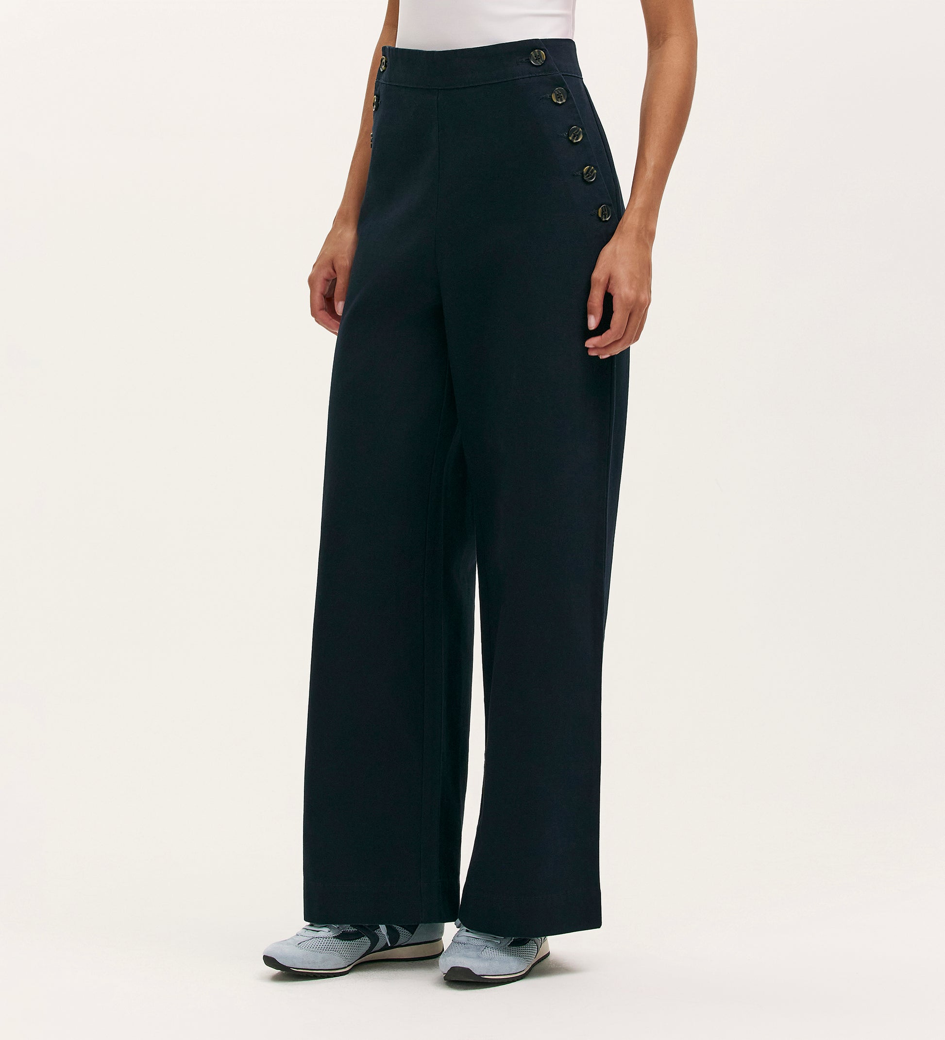 A person in Nellie Navy Canvas Wide Leg Trousers with side button details, a white sleeveless top, and gray sneakers stands against a plain light background—a timeless choice for modern style.