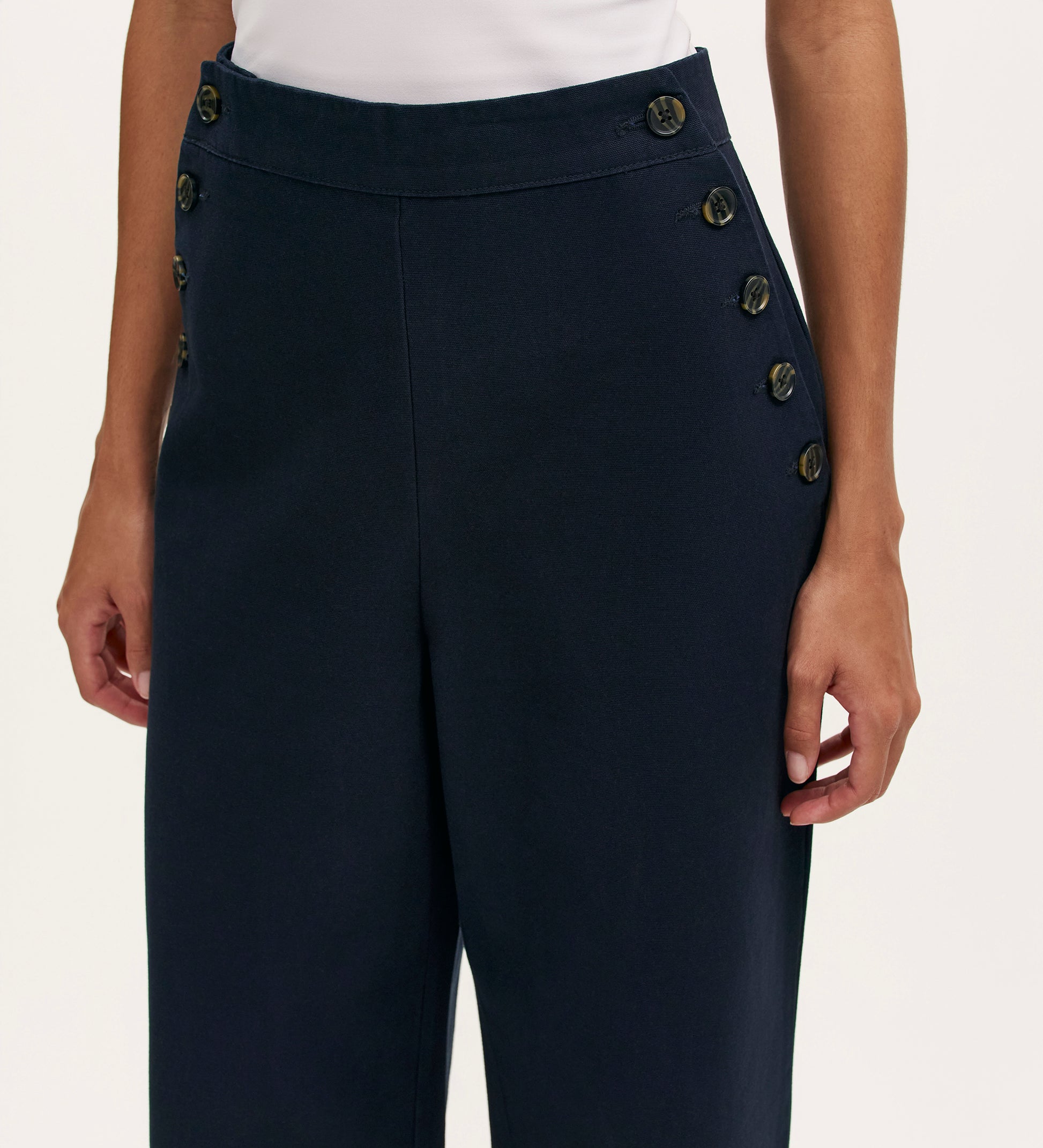 A person wears Nellie Navy Canvas Wide Leg Trousers featuring two rows of black buttons near the pockets, paired with a white top. The image focuses on the waist and upper legs.