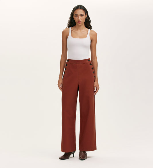 A woman faces forward in a white tank top, brown heeled boots, and Nellie Terracotta Canvas Wide Leg Trousers with side buttons—an effortless wardrobe staple set against a plain light background.