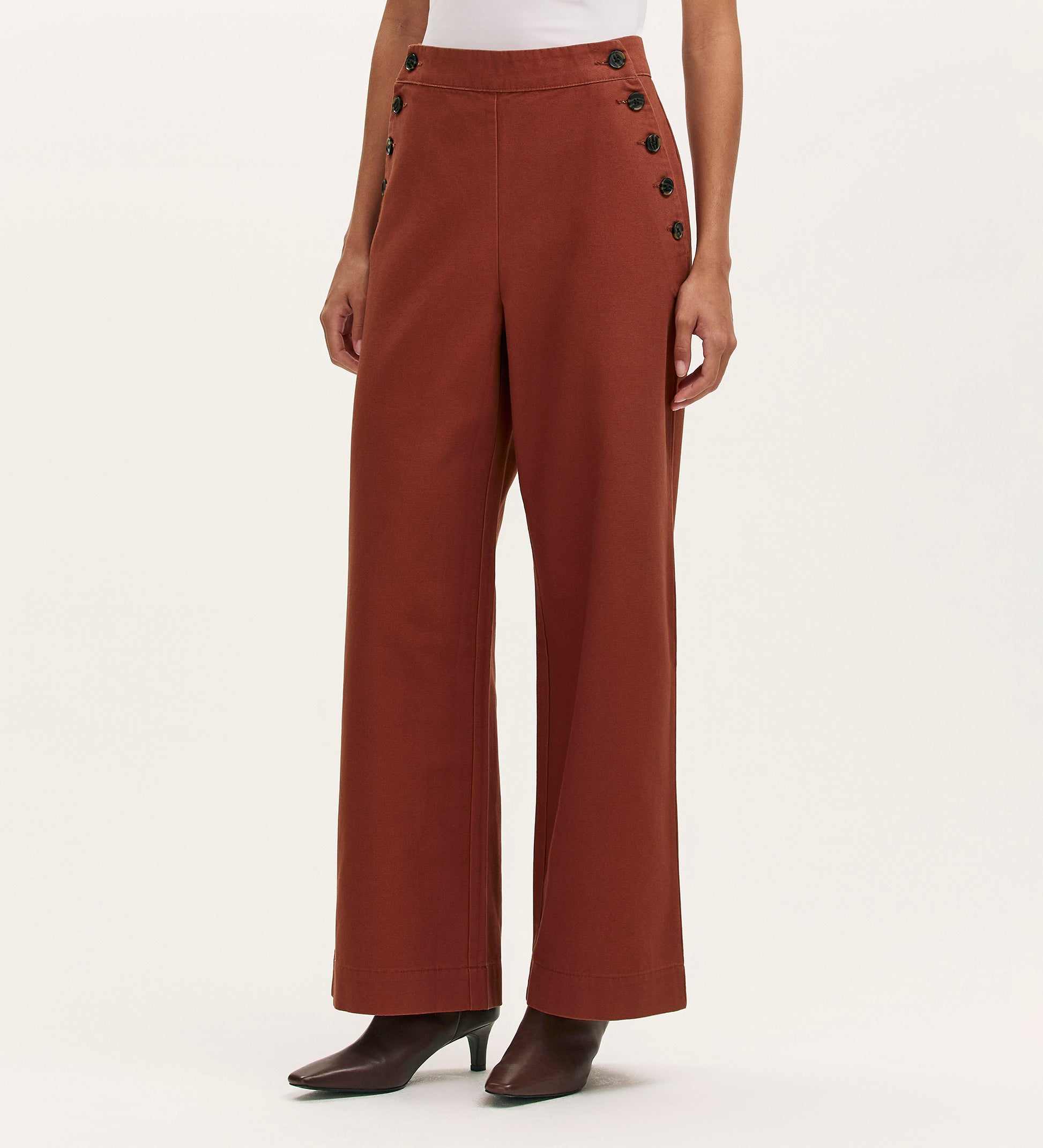 A person wearing Nellie Terracotta Canvas Wide Leg Trousers with black side buttons, paired with a white top and dark brown ankle boots, stands against a plain background.