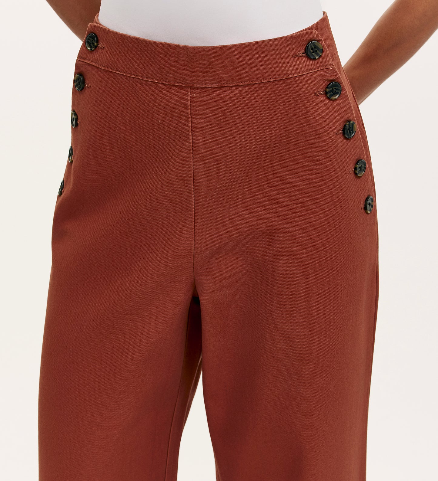 A person models the Nellie Terracotta Canvas Wide Leg Trousers, featuring high-waisted rust fabric and two vertical rows of black buttons on the side panels. The shot shows only the lower torso and upper legs against a plain background.