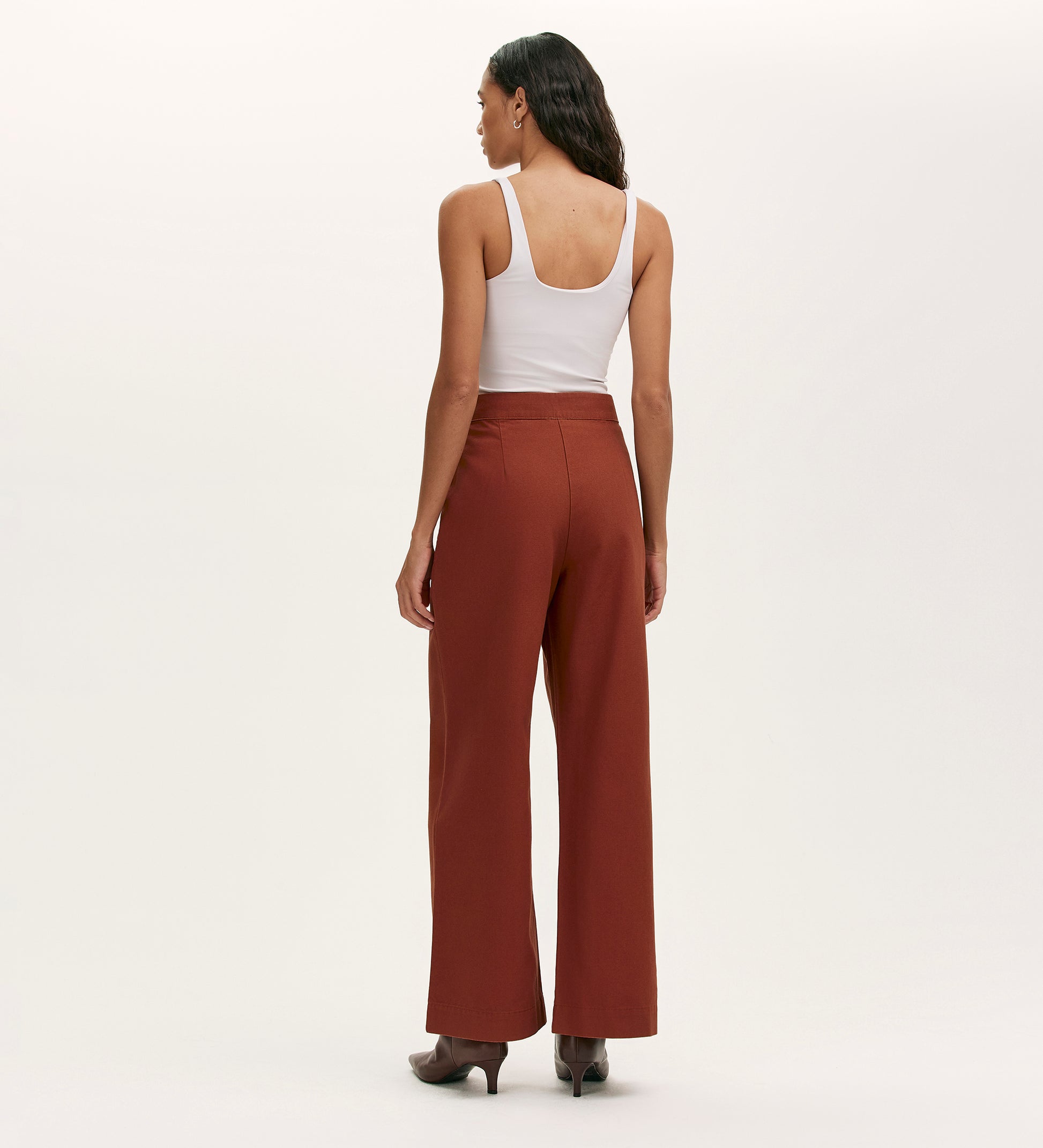 A woman stands facing away, wearing a white sleeveless top, Nellie Terracotta Canvas Wide Leg Trousers, and heeled boots against a plain light background.