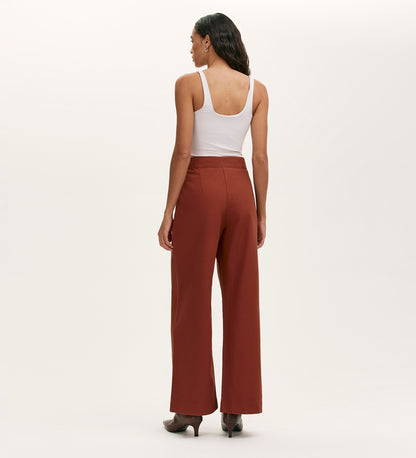 A woman stands facing away, wearing a white sleeveless top, Nellie Terracotta Canvas Wide Leg Trousers, and heeled boots against a plain light background.