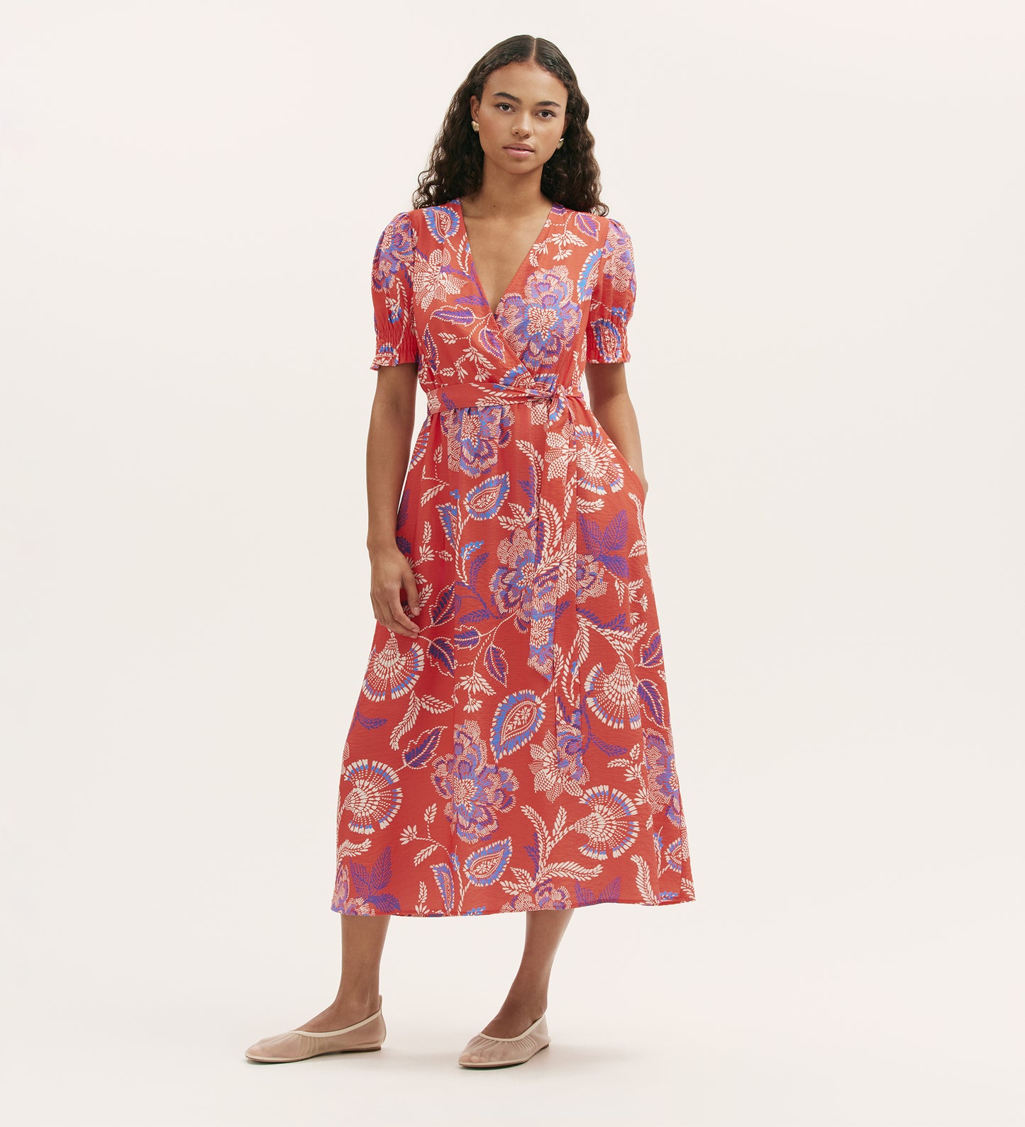 A woman with long, wavy hair wears the Nicoleta Coral Floral Midi Dress, featuring a red floral print, V-neck, wrapped-front bodice, and short sleeves. She pairs it with flat nude shoes, standing against a plain background.