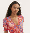 A woman with long, curly dark hair wears the Nicoleta Coral Floral Midi Dress, featuring a red V-neck, blue and white florals, wrapped-front bodice, and shirred waist. She faces the camera with a neutral look against a plain light background.