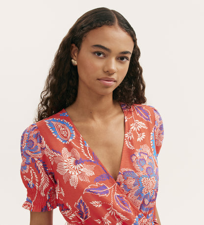 A woman with long, curly dark hair wears the Nicoleta Coral Floral Midi Dress, featuring a red V-neck, blue and white florals, wrapped-front bodice, and shirred waist. She faces the camera with a neutral look against a plain light background.