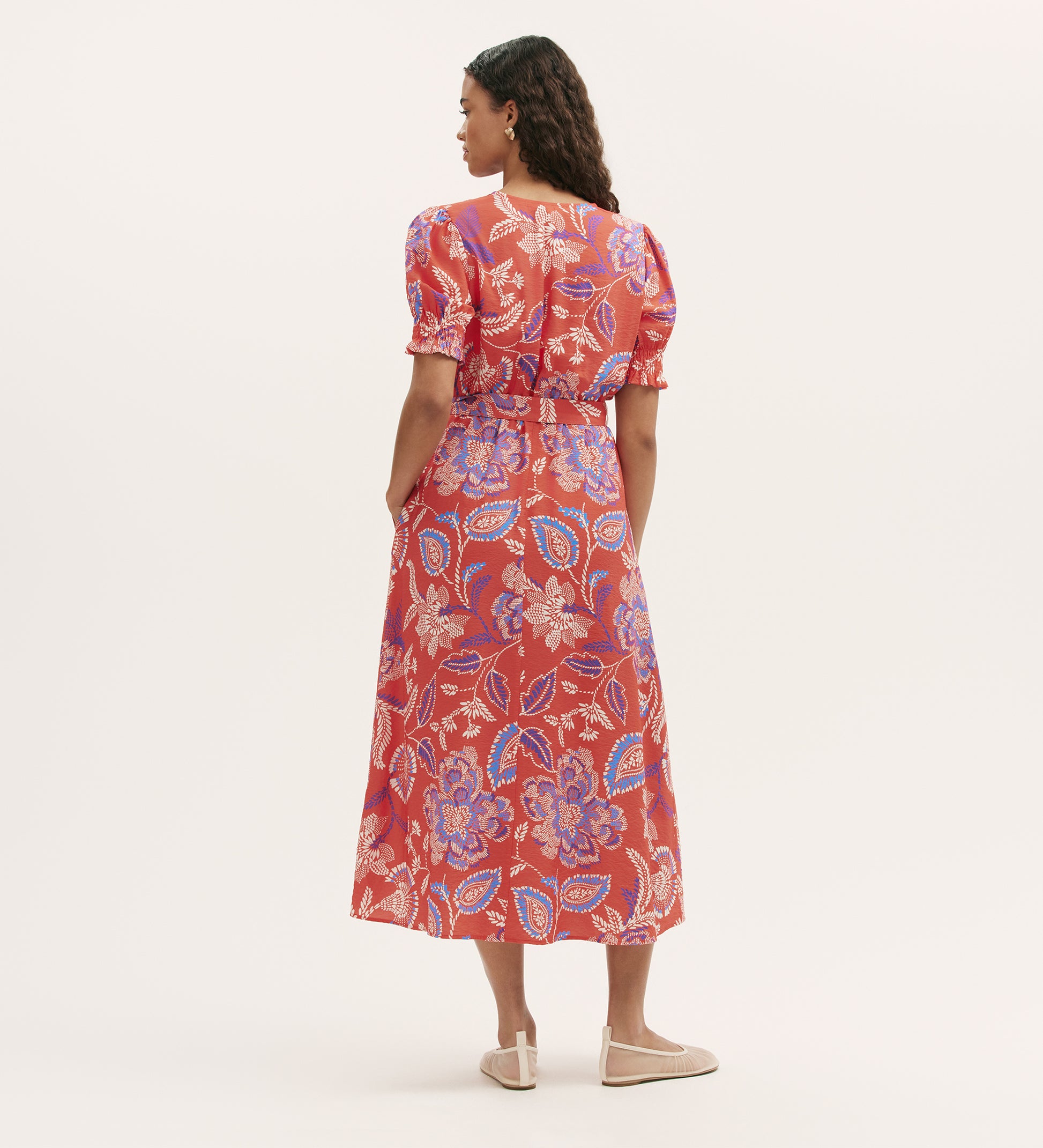 A woman stands with her back to the camera, wearing the Nicoleta Coral Floral Midi Dress—featuring a red floral print, V-neck, puff sleeves, and shirred waist—paired with beige flats against a plain light background.