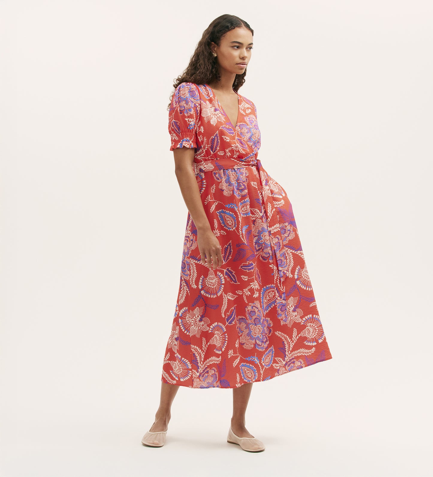 A woman wears the Nicoleta Coral Floral Midi Dress, featuring a red floral pattern, V-neck, short sleeves, wrap-front bodice, and tie at the shirred waist. She has long wavy hair and light flat shoes against a plain background.