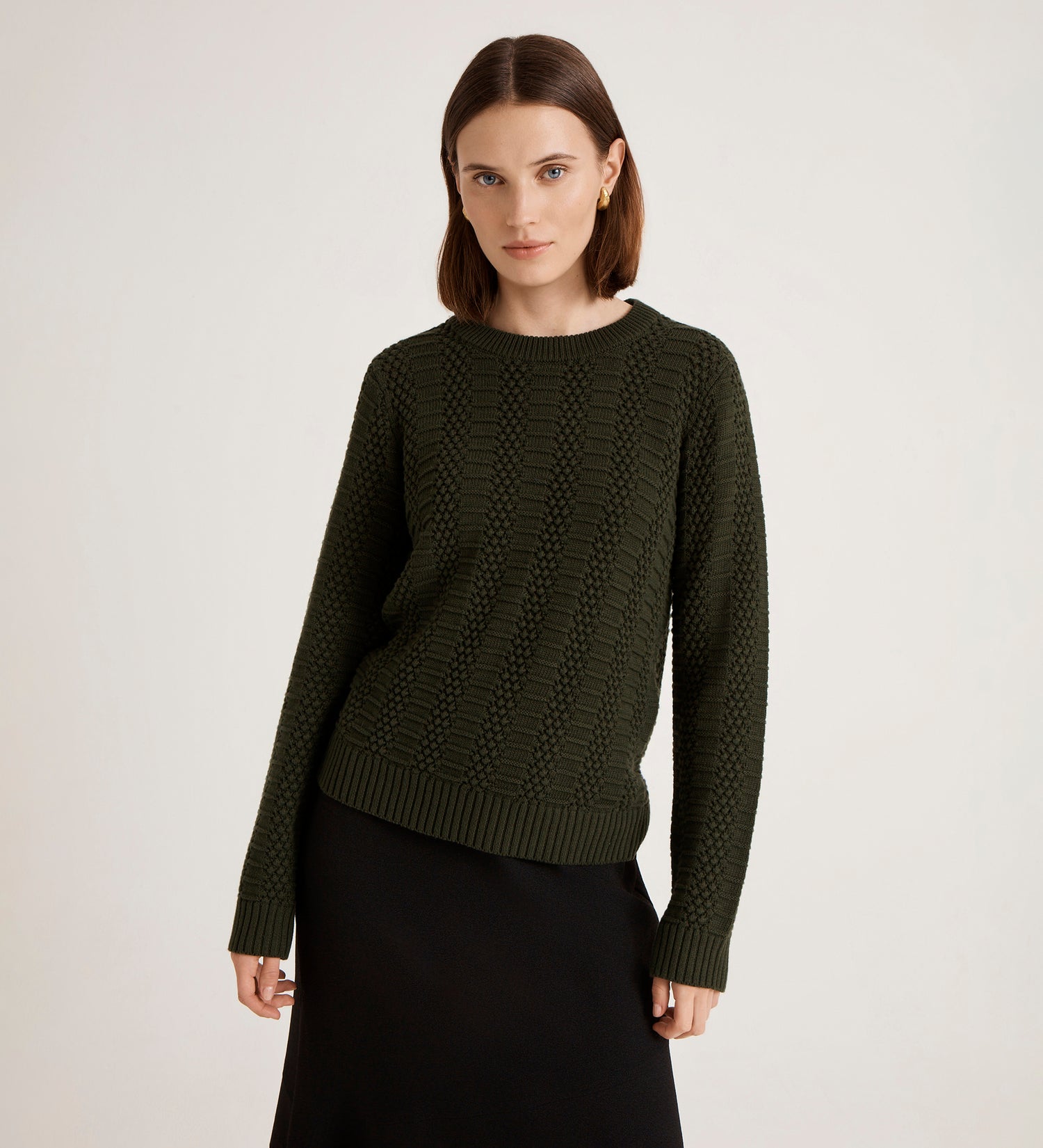 A woman with straight brown hair models the Nova Green Knitted Jumper, paired with a black skirt against a plain light background. With a relaxed pose and neutral expression, this textured piece is a timeless staple for any women’s wardrobe.