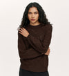 A woman with long, curly dark hair wears the Odette Mole Knitted Jumper in dark brown, paired with black pants, standing against a plain light background with her left arm crossing her body to gently touch her right shoulder.