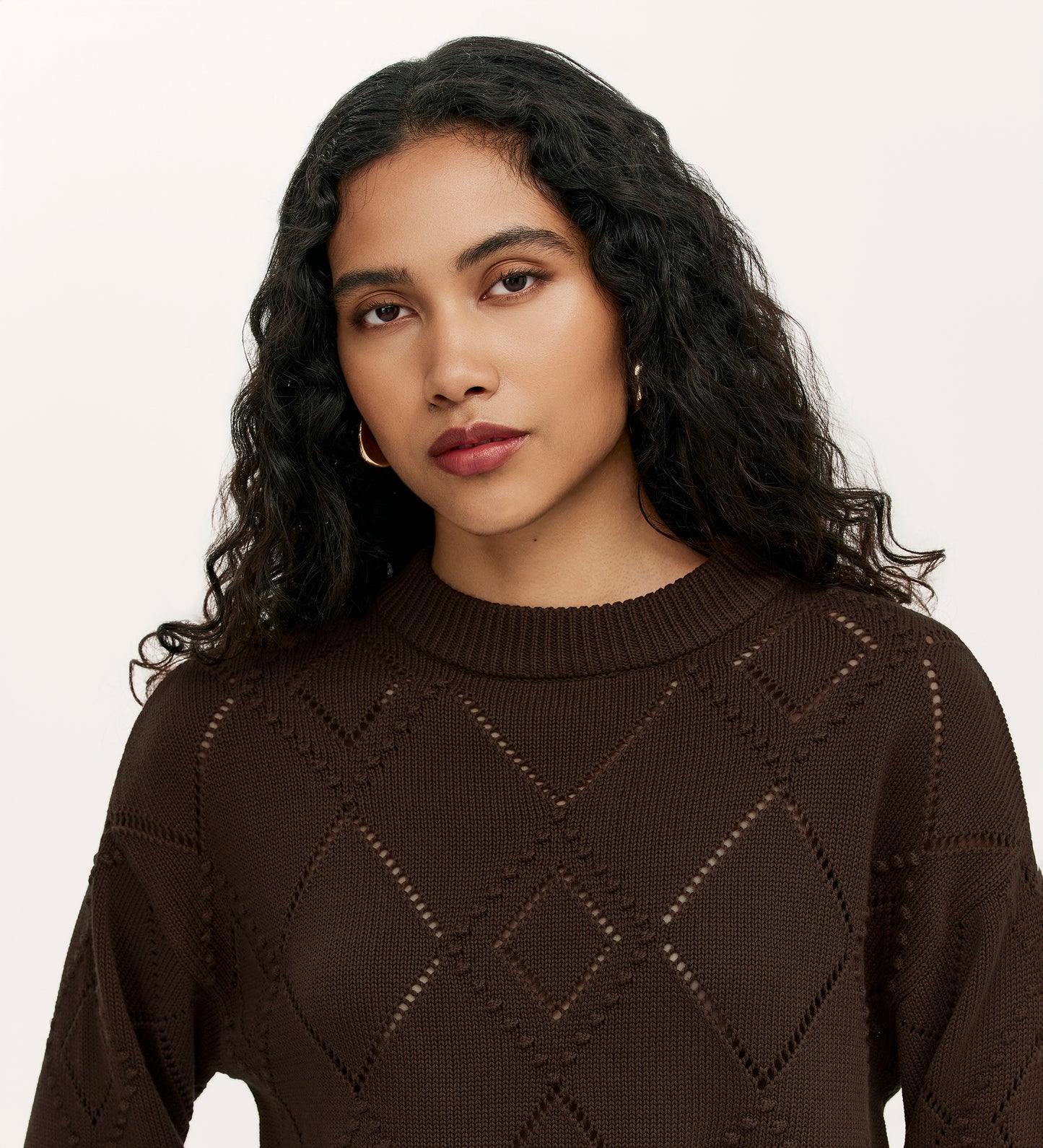 A woman with long, curly dark hair and gold hoop earrings wears the Odette Mole Knitted Jumper, standing against a plain light background and looking directly at the camera with a neutral expression.