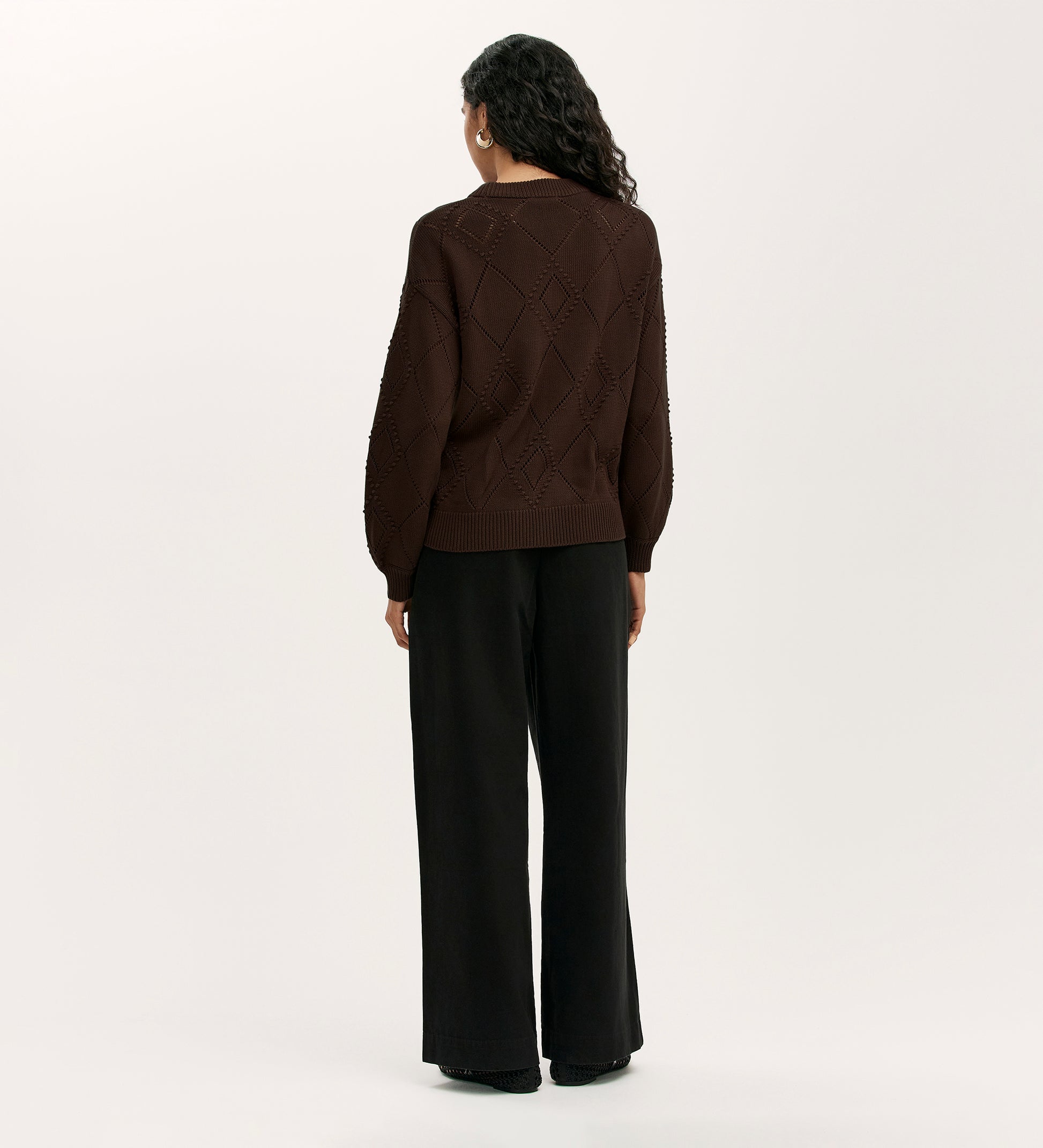 A person with long curly hair stands facing away, wearing the Odette Mole Knitted Jumper and loose black pants against a plain light background.