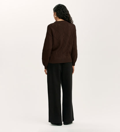 A person with long curly hair stands facing away, wearing the Odette Mole Knitted Jumper and loose black pants against a plain light background.