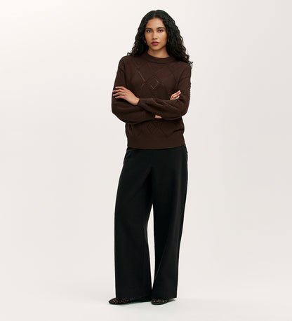 A person with long, curly dark hair wears the Odette Mole Knitted Jumper and loose black pants, standing against a plain, light background with arms loosely crossed.