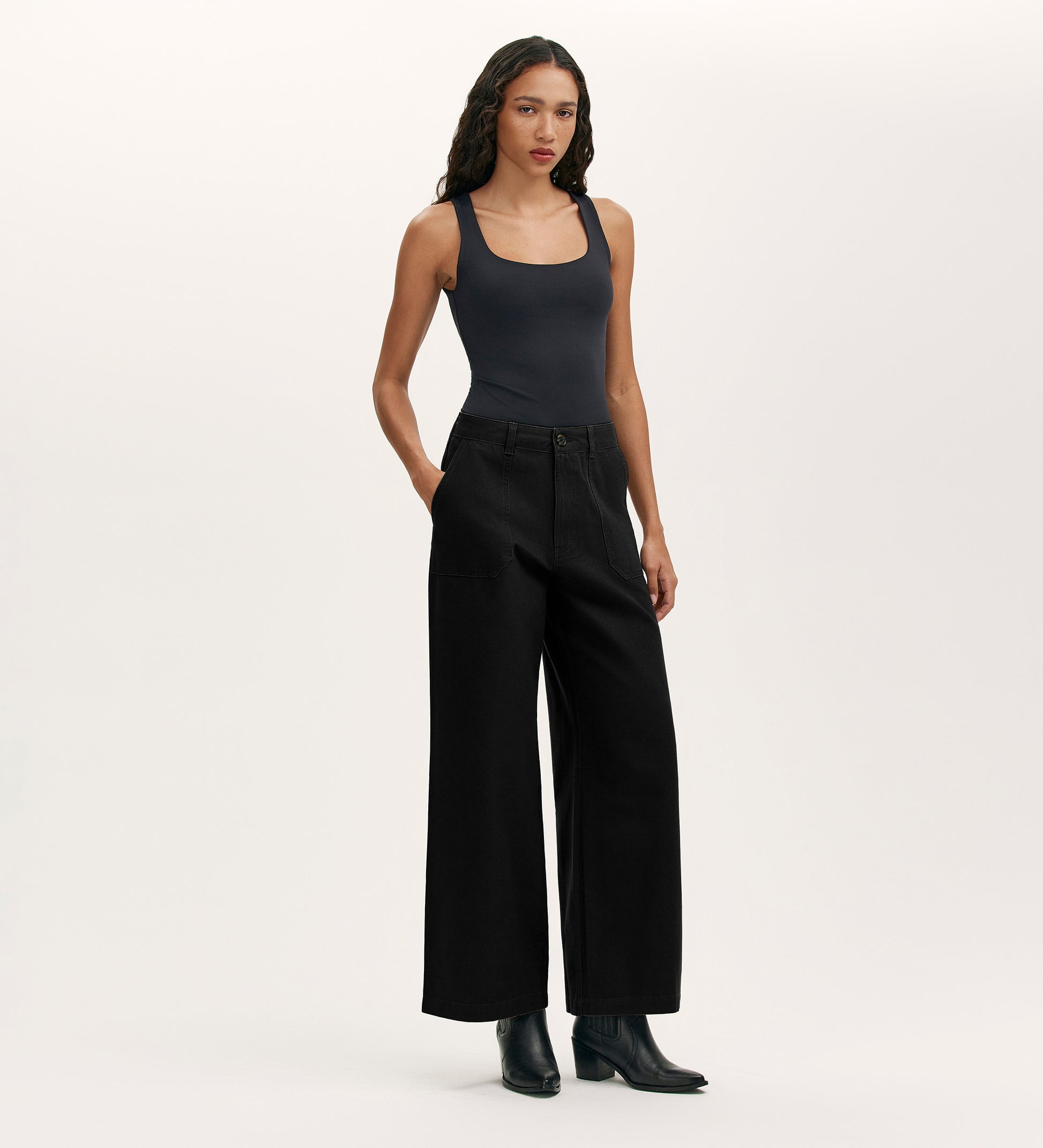 A person stands against a plain background, wearing Olive Black Canvas Wide Leg Trousers with a fitted black tank top and black ankle boots. Their hands are in their pockets, and long wavy hair completes this elevated casual look.