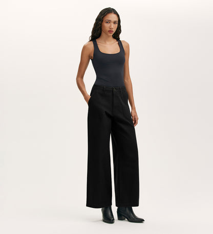 A person stands against a plain background, wearing Olive Black Canvas Wide Leg Trousers with a fitted black tank top and black ankle boots. Their hands are in their pockets, and long wavy hair completes this elevated casual look.