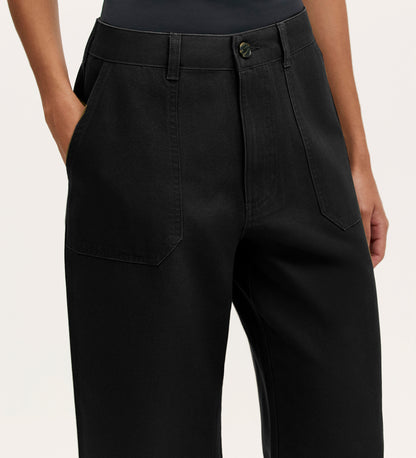 A person wears Olive Black Canvas Wide Leg Trousers featuring a high waist, large front pockets, and button closure, standing hands-in-pockets against a plain background—an effortless, elevated casual look.