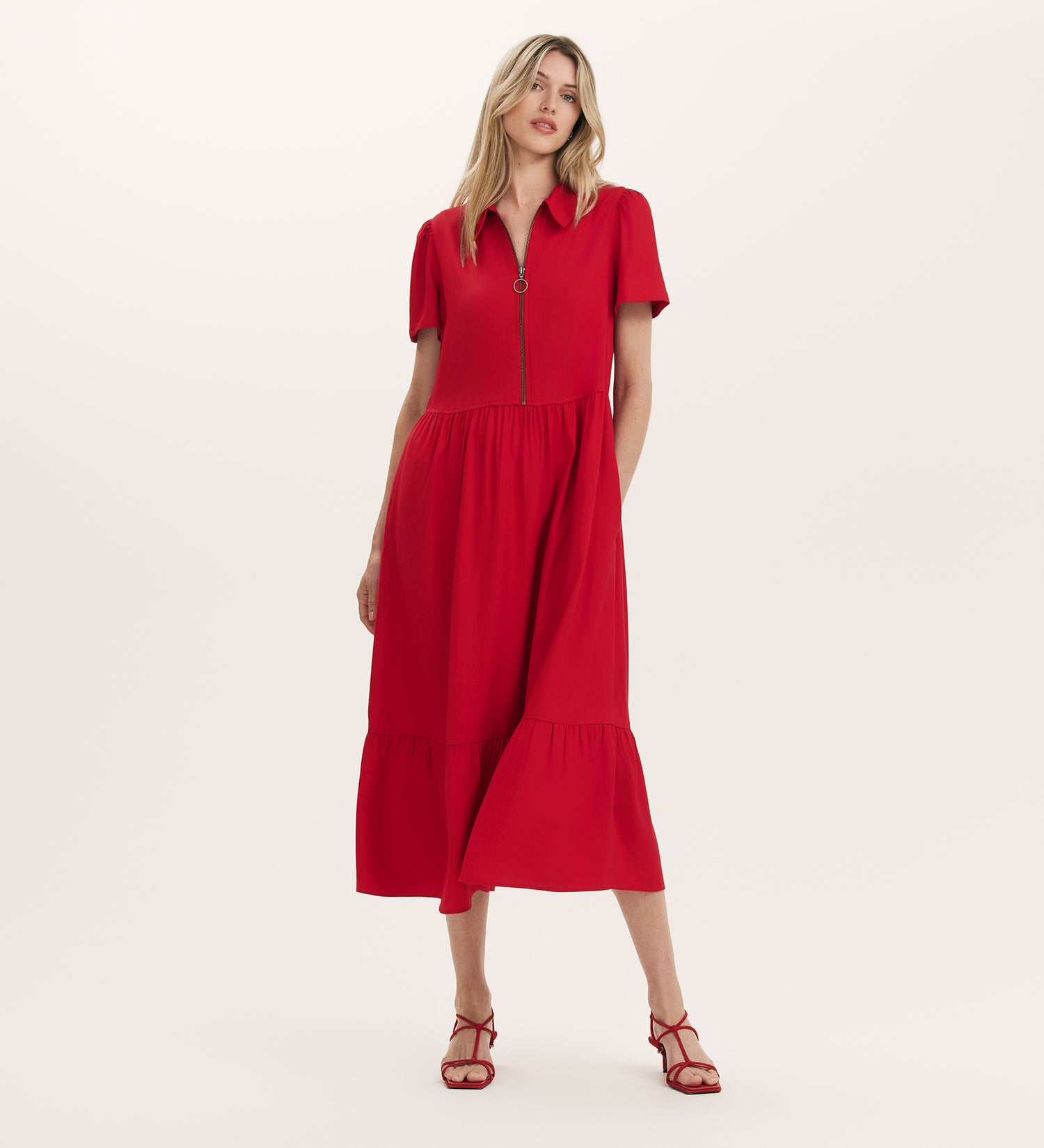 A woman stands against a plain light background wearing the Orion Red Zip Front Midi Shirt Dress with short sleeves, a zipper neckline, and tiered hemline, paired with matching red strappy sandals.