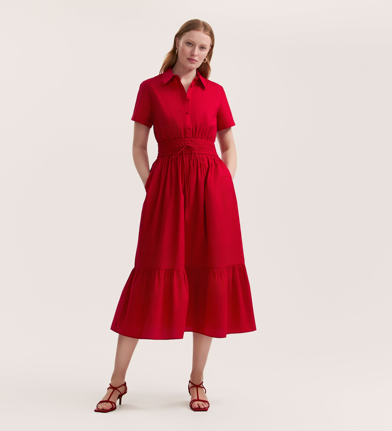A woman wears the Penny Red Midi Dress, featuring short sleeves, an elasticated waist, and a tiered skirt, paired with red strappy heels against a plain background.