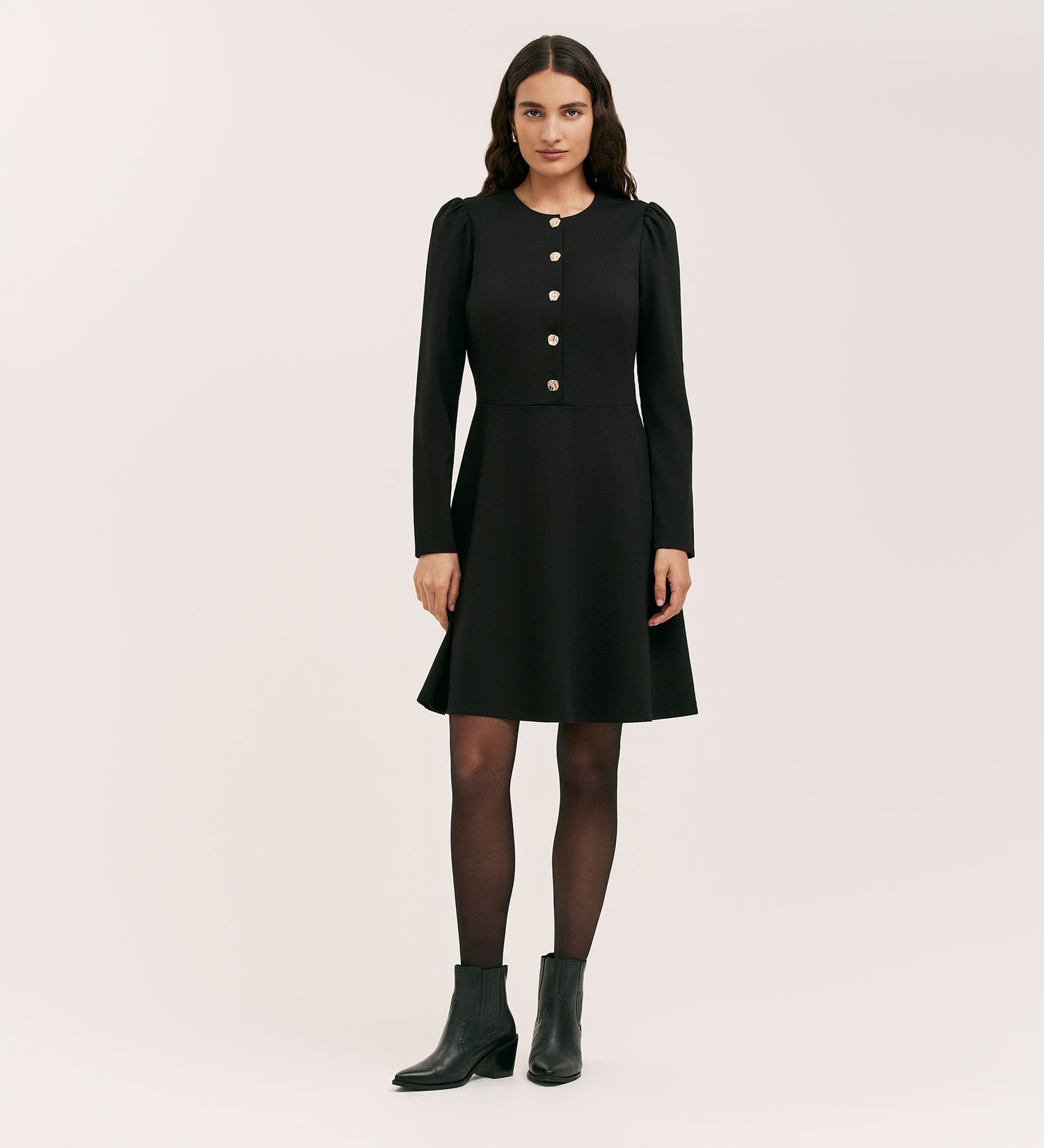 A woman with long dark hair wears the Rae Black Ponte Jersey Short Dress, styled with black tights and black ankle boots. She stands facing forward against a plain light background.