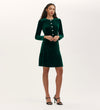 A woman poses against a plain light background wearing the Rae Bottle Green Velvet Short Dress, knee-length with long sleeves and large front buttons, paired with black heeled sandals.