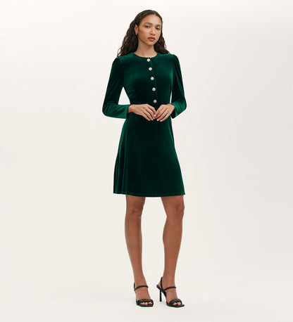 A woman poses against a plain light background wearing the Rae Bottle Green Velvet Short Dress, knee-length with long sleeves and large front buttons, paired with black heeled sandals.