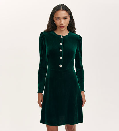 A woman stands against a plain light background wearing the Rae Bottle Green Velvet Short Dress, featuring long sleeves, a round neckline, and decorative buttons down the front.