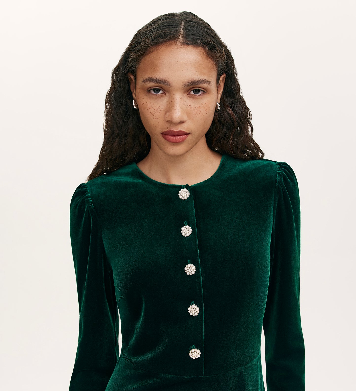 A woman with long curly hair, light brown skin, and freckles wears the Rae Bottle Green Velvet Short Dress against a plain light background, looking directly at the camera.