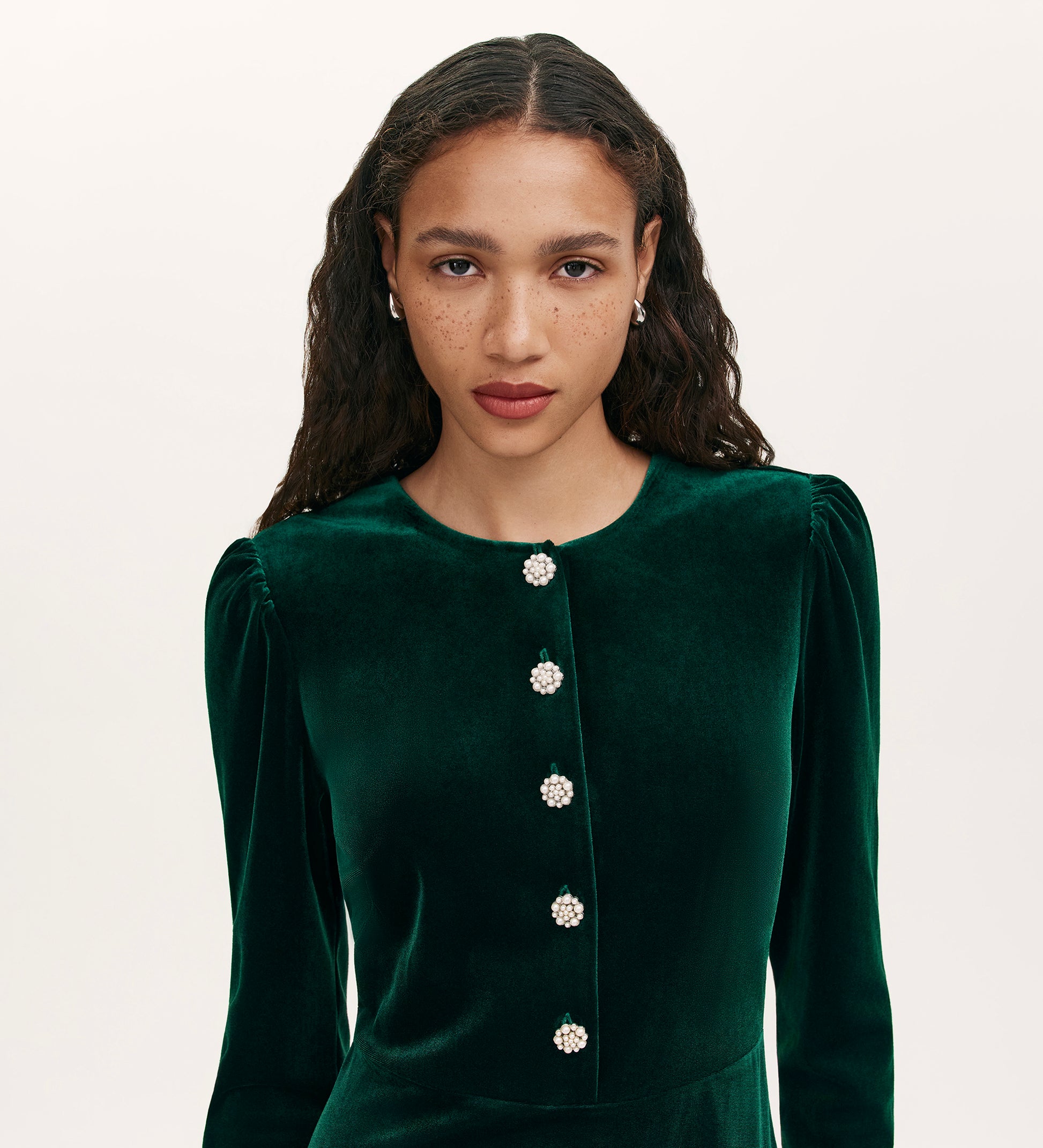 A woman with long curly hair, light brown skin, and freckles wears the Rae Bottle Green Velvet Short Dress against a plain light background, looking directly at the camera.