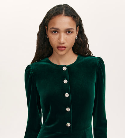 A woman with long curly hair, light brown skin, and freckles wears the Rae Bottle Green Velvet Short Dress against a plain light background, looking directly at the camera.