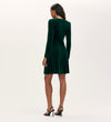 A woman faces away, wearing the Rae Bottle Green Velvet Short Dress, a knee-length, long sleeve A-line party dress. She pairs it with black high-heeled sandals against a plain light background.