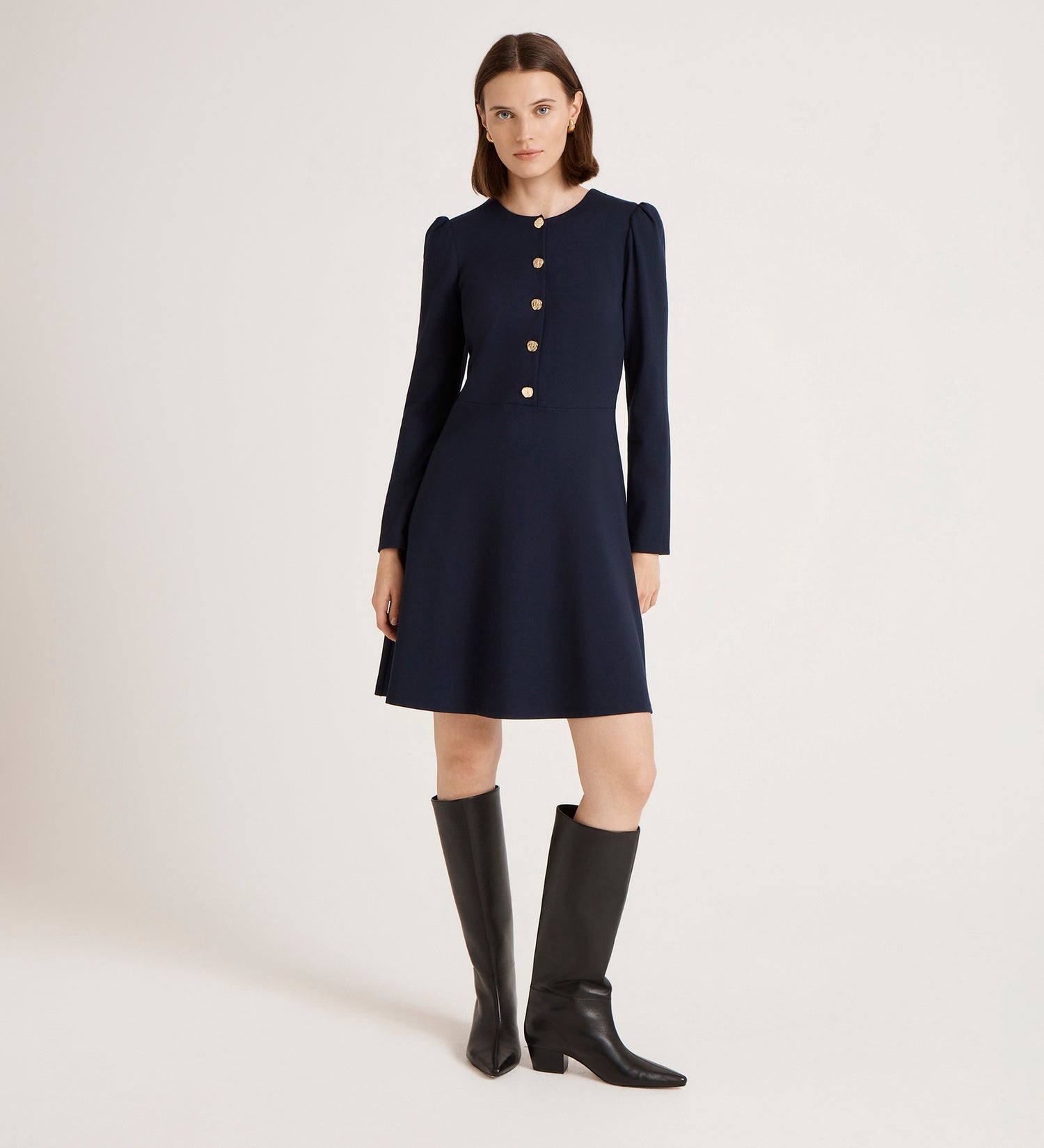 A woman stands against a plain background, wearing the Rae Navy Ponte Jersey Short Dress with gold buttons down the front and black knee-high boots.