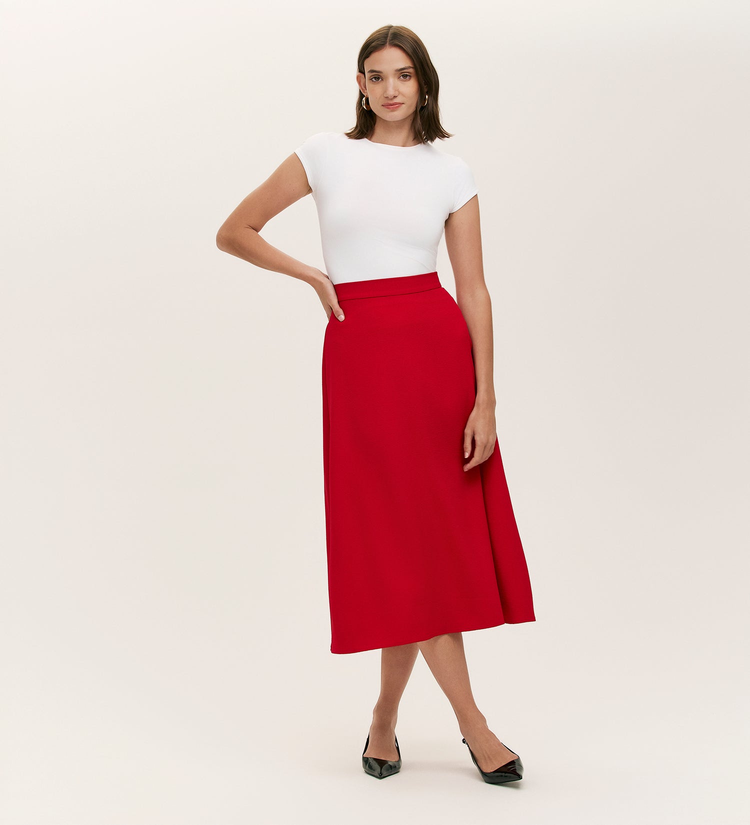 A woman poses with a hand on her hip, exuding timeless appeal in the Ramona Red Rich Crepe Midi Skirt, styled with a white fitted short-sleeve top and black pointed-toe heels against a plain background.