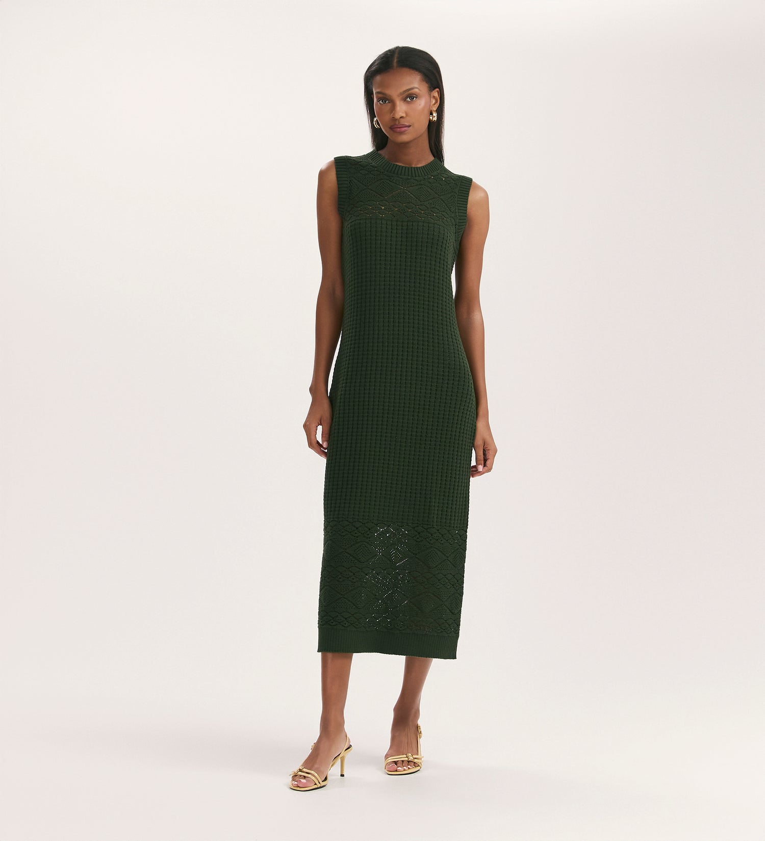 A woman with straight hair and gold earrings wears the Ravi Khaki Sleeveless Knitted Midi Dress, featuring a relaxed fit and subtle patterns. She pairs it with yellow open-toe heels against a plain light background.