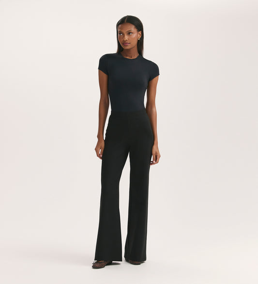 A woman stands against a plain white background, wearing a fitted black short-sleeve top and Reyna Black Ponte Jersey Wide Leg Trousers, paired with black pointed-toe shoes.