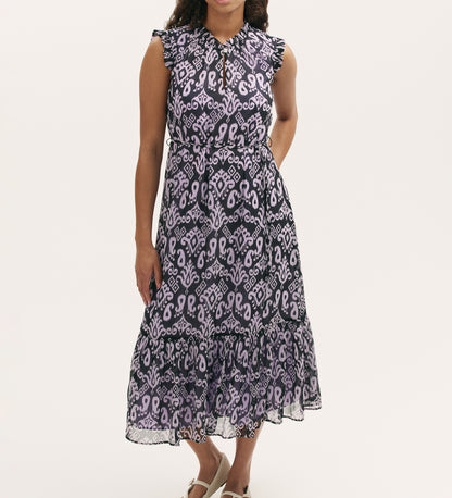 A person wears the Rosalind Lilac Ikat Chiffon Sleeveless Midi Dress, featuring a purple and black abstract pattern and a shirred waist, standing with arms relaxed against a plain light background.