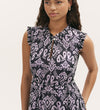 A woman stands with one hand in her pocket, wearing the Rosalind Lilac Ikat Chiffon Sleeveless Midi Dress featuring a purple and black abstract print, ruffled shoulders, shirred waist, keyhole neckline, and tiered skirt.
