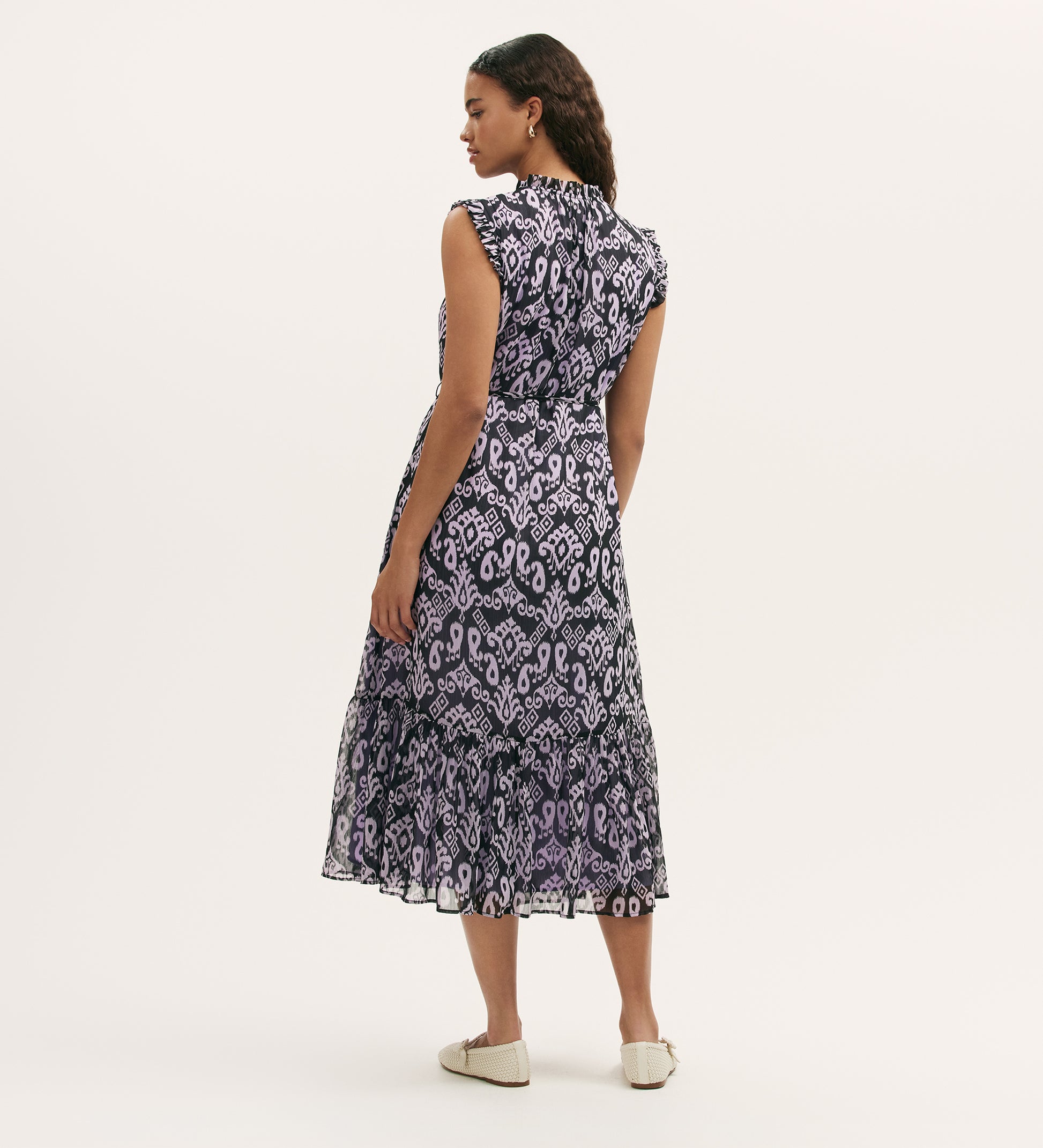 A woman stands facing away, wearing the Rosalind Lilac Ikat Chiffon Sleeveless Midi Dress with a tiered skirt and white flats against a plain, light background.