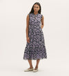 A woman with long dark hair stands against a plain background, hands behind her back, wearing the Rosalind Lilac Ikat Chiffon Sleeveless Midi Dress with a tiered skirt and ruffled hem, paired with white flat shoes.