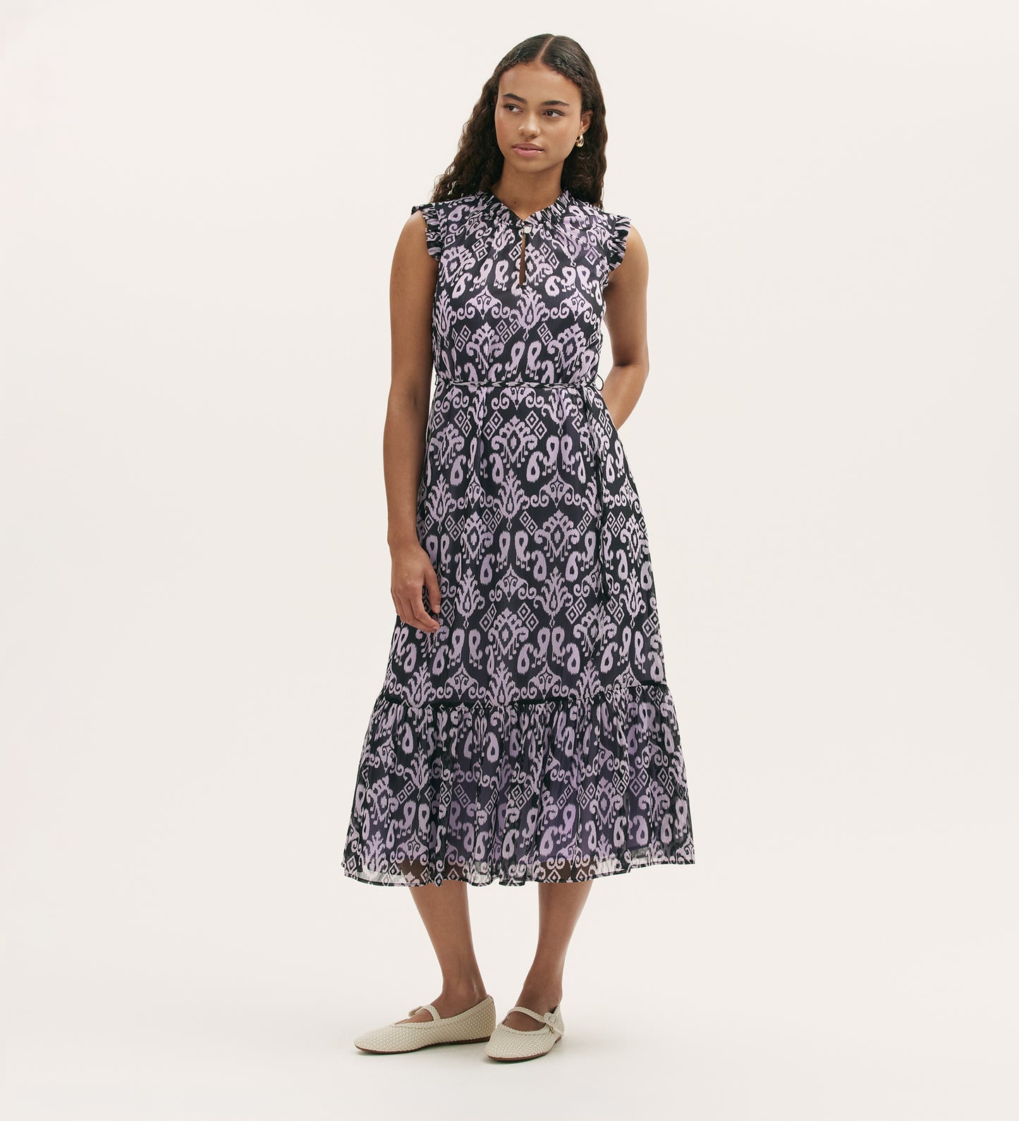A woman with long dark hair stands against a plain background, hands behind her back, wearing the Rosalind Lilac Ikat Chiffon Sleeveless Midi Dress with a tiered skirt and ruffled hem, paired with white flat shoes.