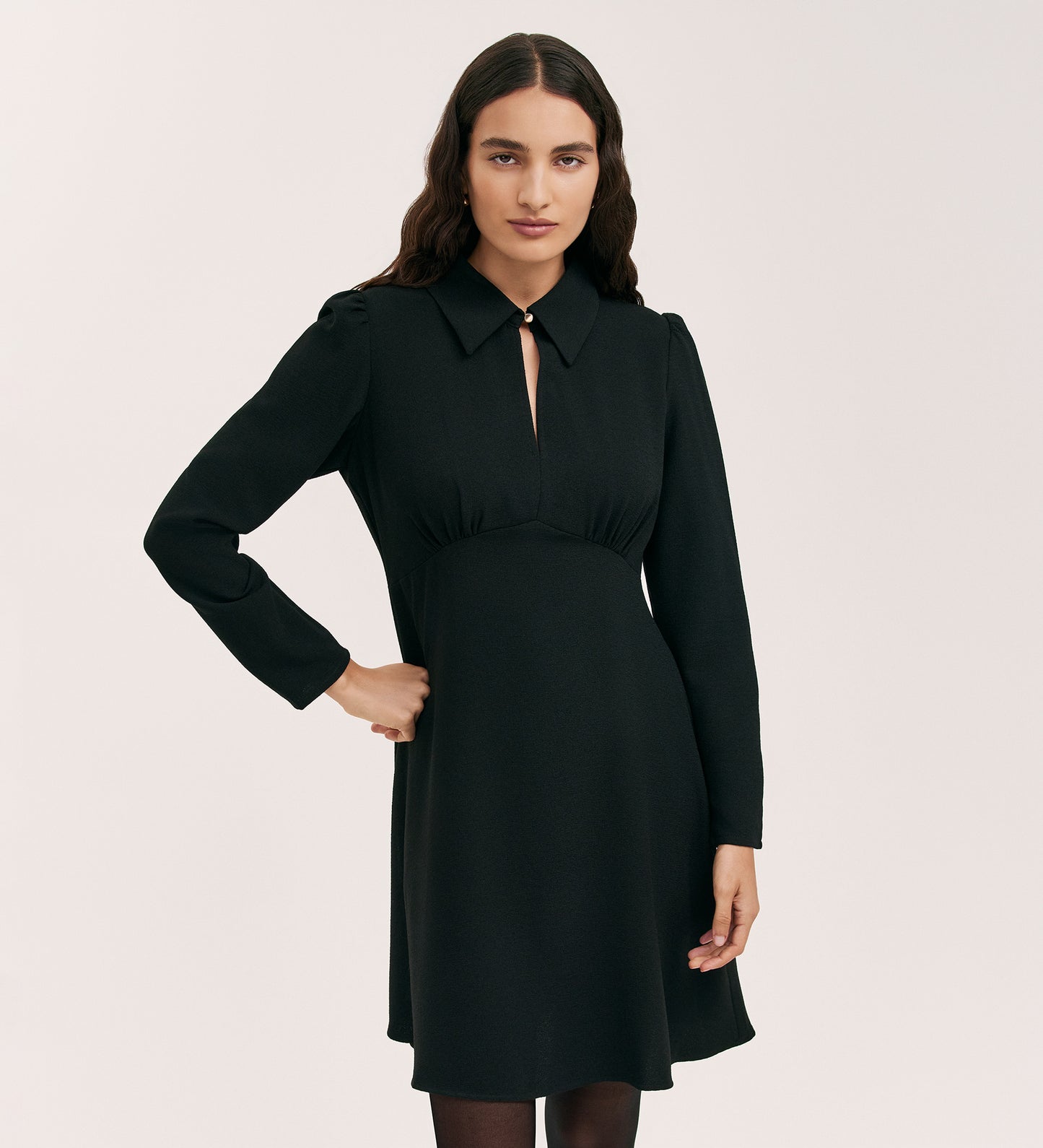A woman with long, wavy brown hair models the Sabine Black Crepe Short Dress, featuring a fitted cut, long sleeves, knee-length hem, front keyhole detail, and pointed collar, set against a plain light background.