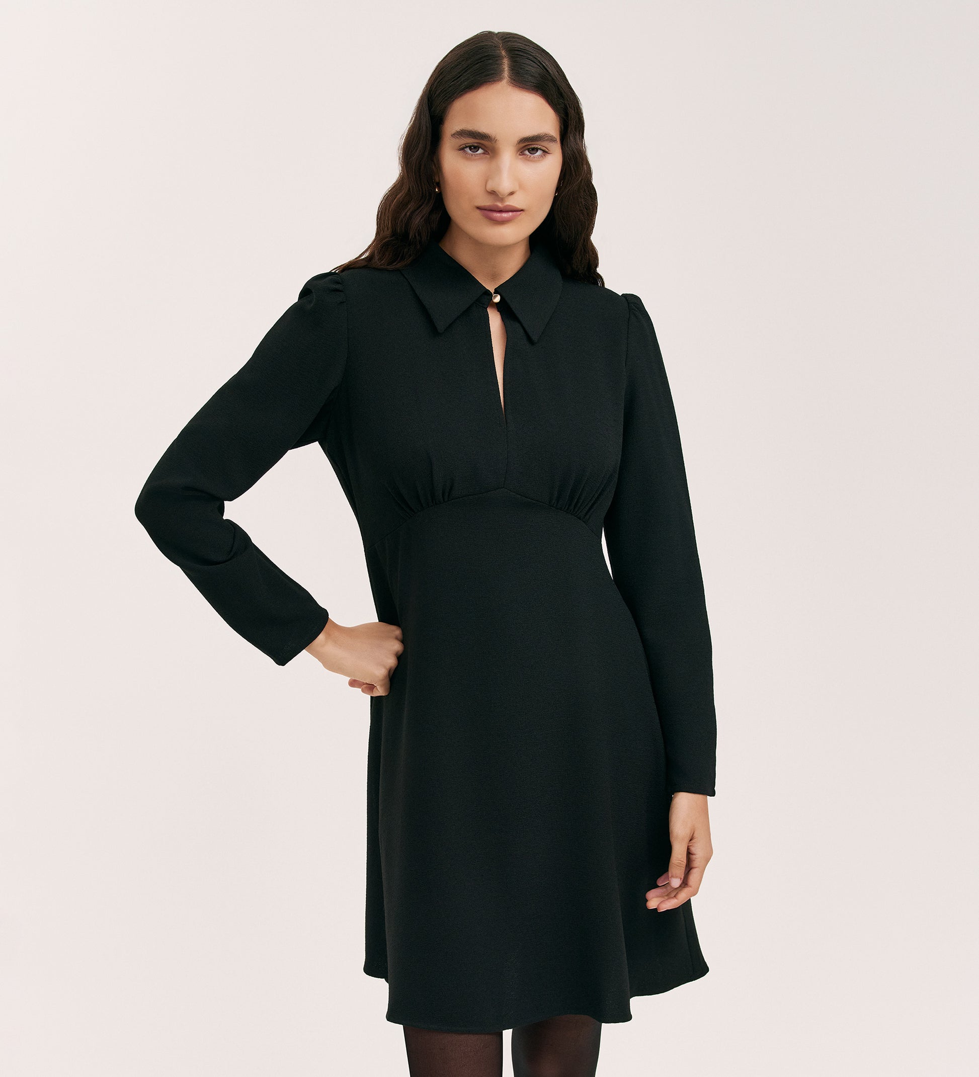 A woman with long, wavy brown hair models the Sabine Black Crepe Short Dress, featuring a fitted cut, long sleeves, knee-length hem, front keyhole detail, and pointed collar, set against a plain light background.